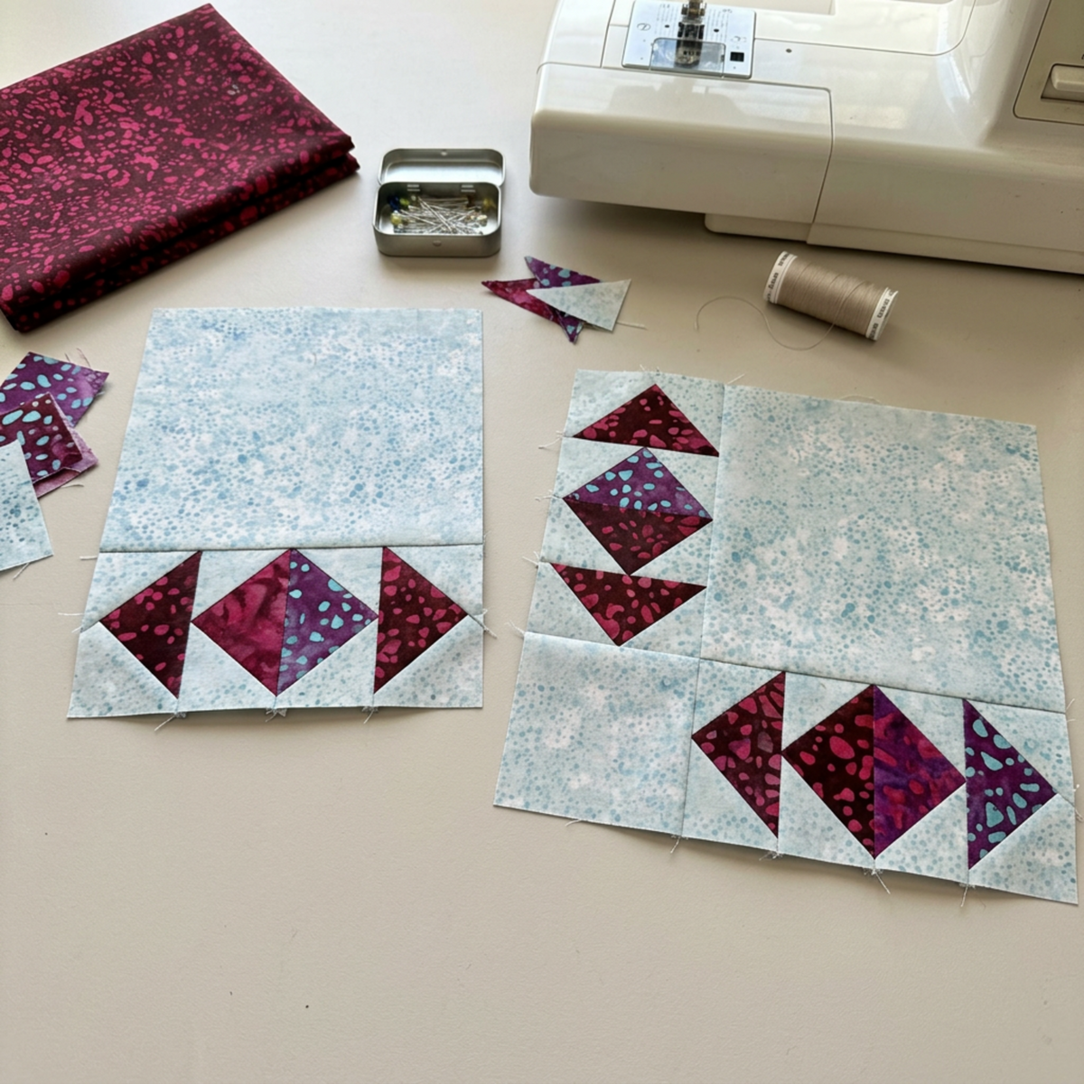 Two unfinished quilt blocks with burgundy and purple triangle-and-diamond units along the edges of large pale background areas, beside folded Mahogany fabric, pins, thread, and a sewing machine. Quilts shown for inspiration feature the Geese Over Galveston Bay Quilt Pattern by The Quilt Rambler, available at IraRobi Quilts.