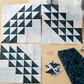 Three unfinished ocean-wave quilt blocks in dark teal and pale blue-gray batiks on a sewing table with folded Smoky Teal fabric, scraps, pins, and thread. Quilts shown for inspiration feature the Kelley Quilt Pattern by Edyta Sitar of Laundry Basket Quilts, available at IraRobi Quilts.