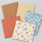 Five folded Lazy Afternoon fabrics arranged in a fan, showing bronze dots, gold geometric lines, blue plus signs, coral lines, and pastel yarn-ball prints.