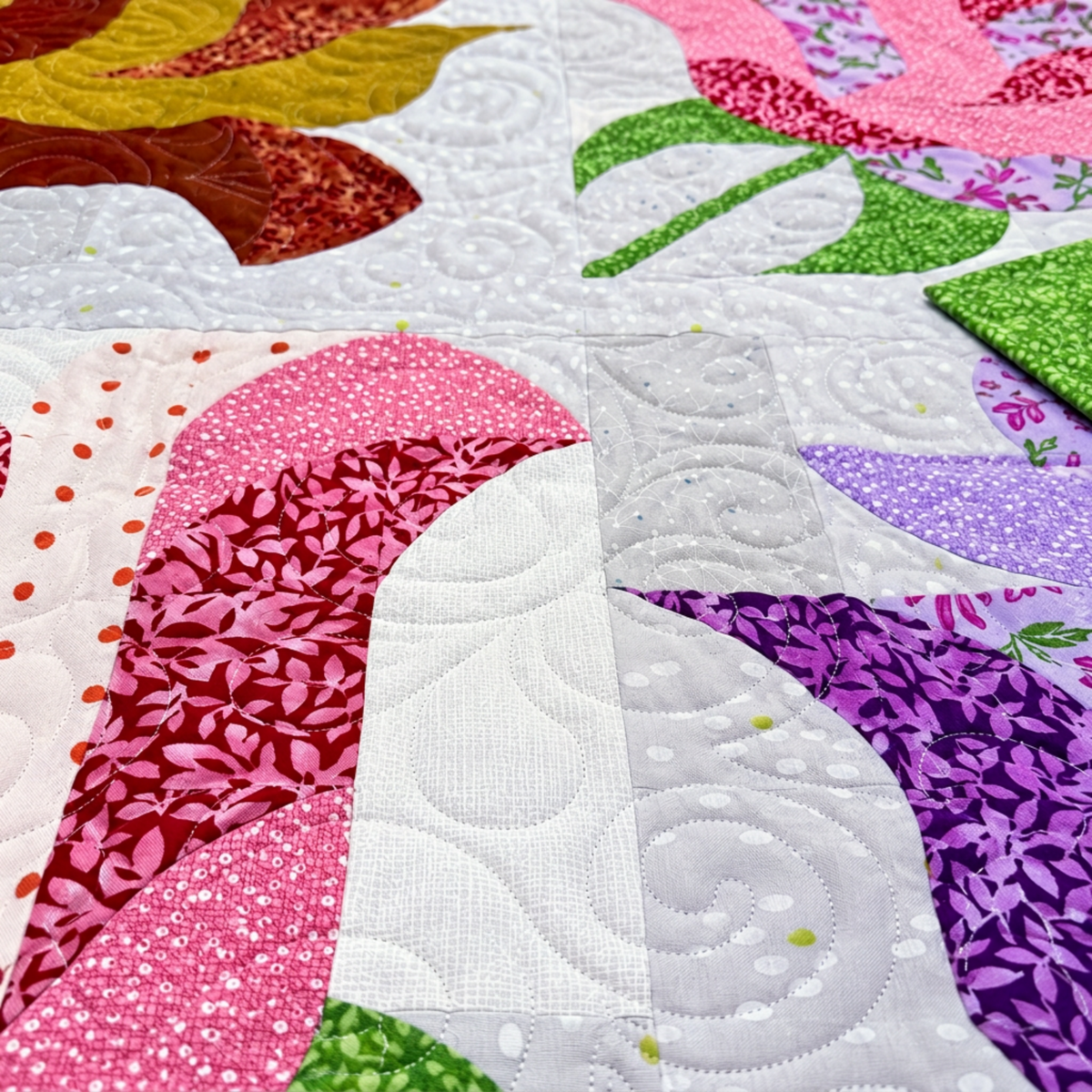 Close-up of Rose Garden quilt showing pink and purple curved patchwork, green leaves, pale printed fabrics, and spiral quilting.