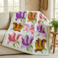 Rose Garden floral quilt draped over a cream sofa beside a wooden table with a potted fern in a sunlit room.