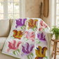 Rose Garden floral quilt draped over a cream sofa beside a wooden table with a potted fern in a sunlit room.