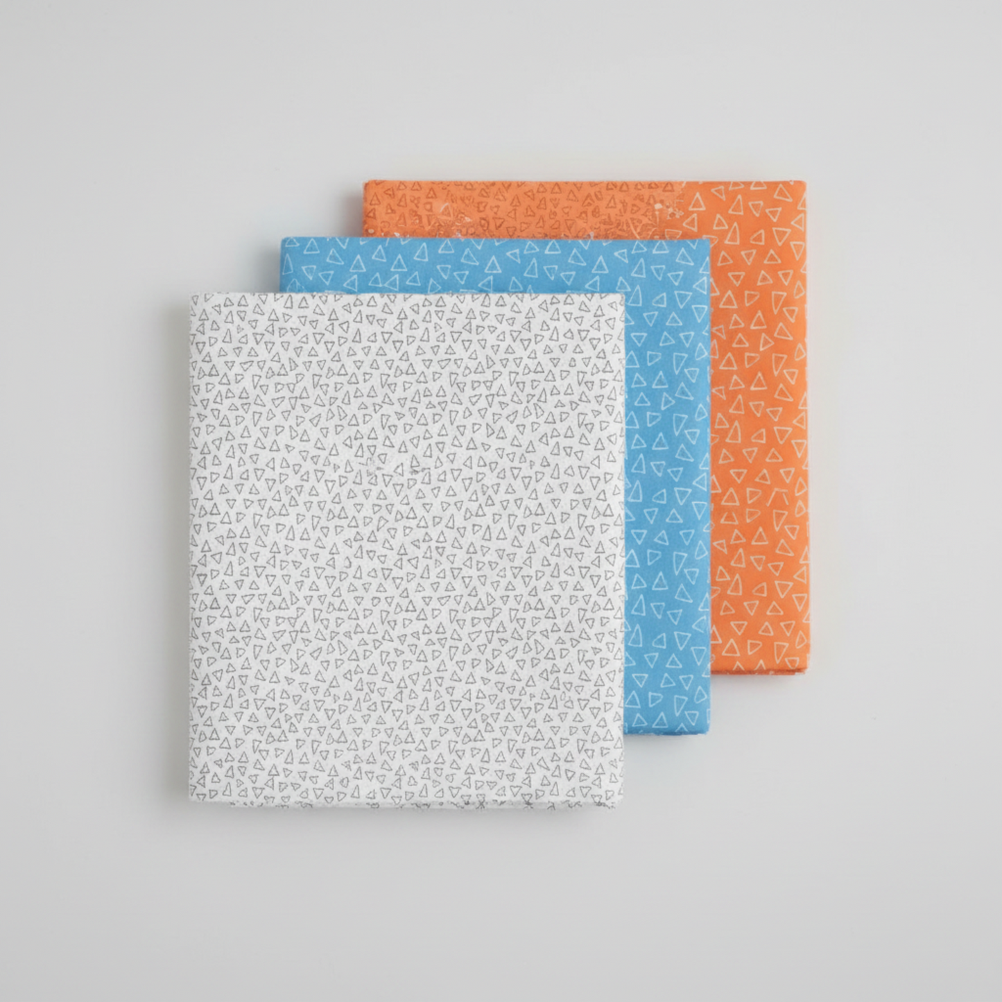 Safari Fun Floating Triangles quilting fabric in three colorways – white with black outlines, cornflower blue with white outlines, and orange with white outlines, folded and stacked on light grey background