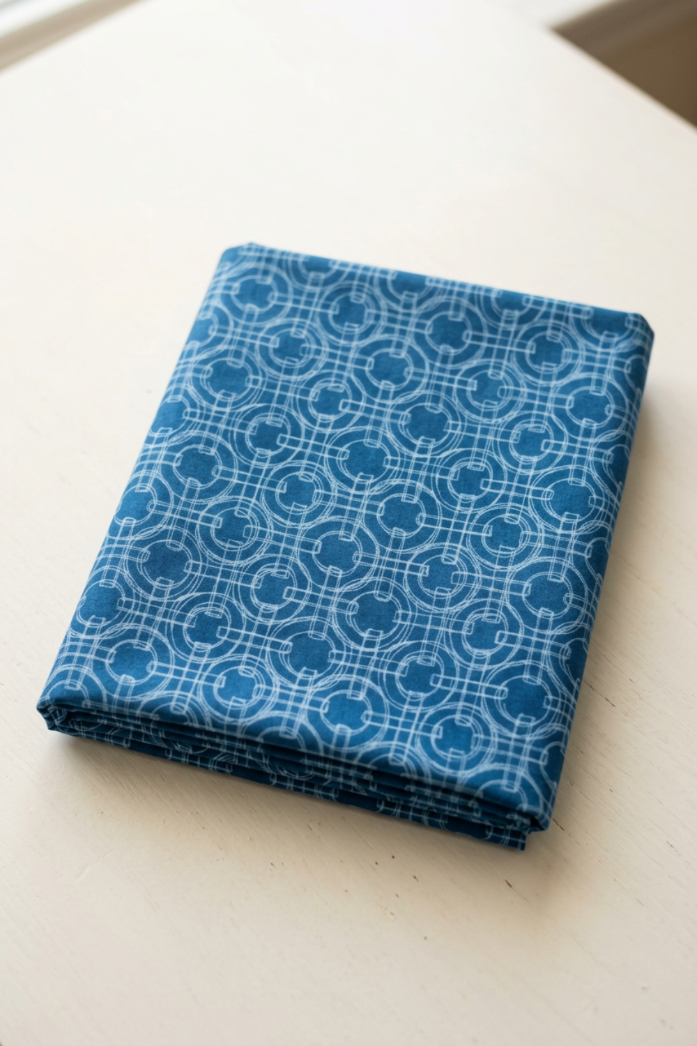 Serena Shores Coastal fabric by Robin Pickens for Moda Fabrics, neatly folded on a light tabletop with pale blue linked rings and intersecting lines on deep blue cotton.