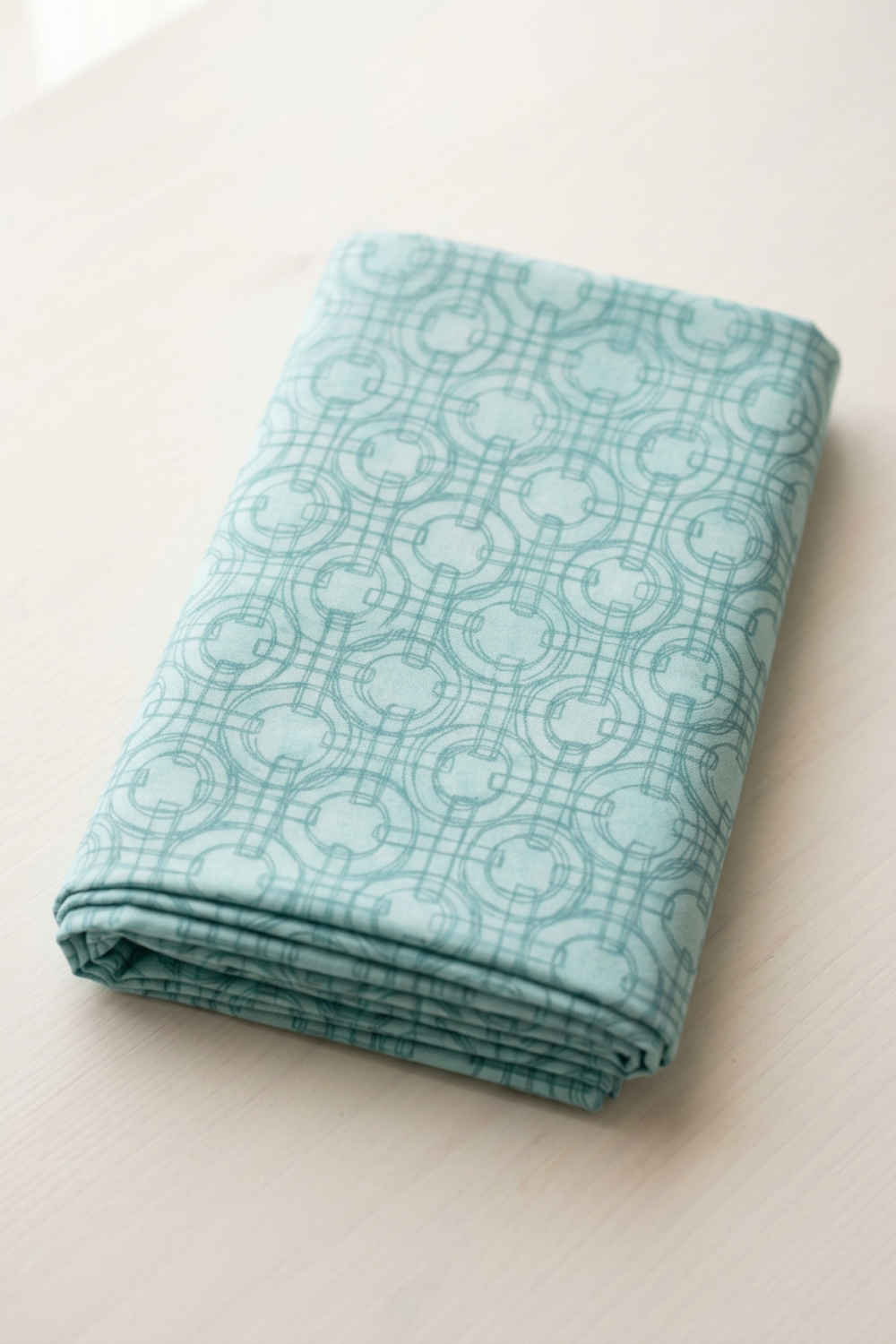 Serena Shores Mist fabric by Robin Pickens for Moda Fabrics, neatly folded on a cream tabletop with muted teal linked rings and intersecting lines on pale aqua cotton.