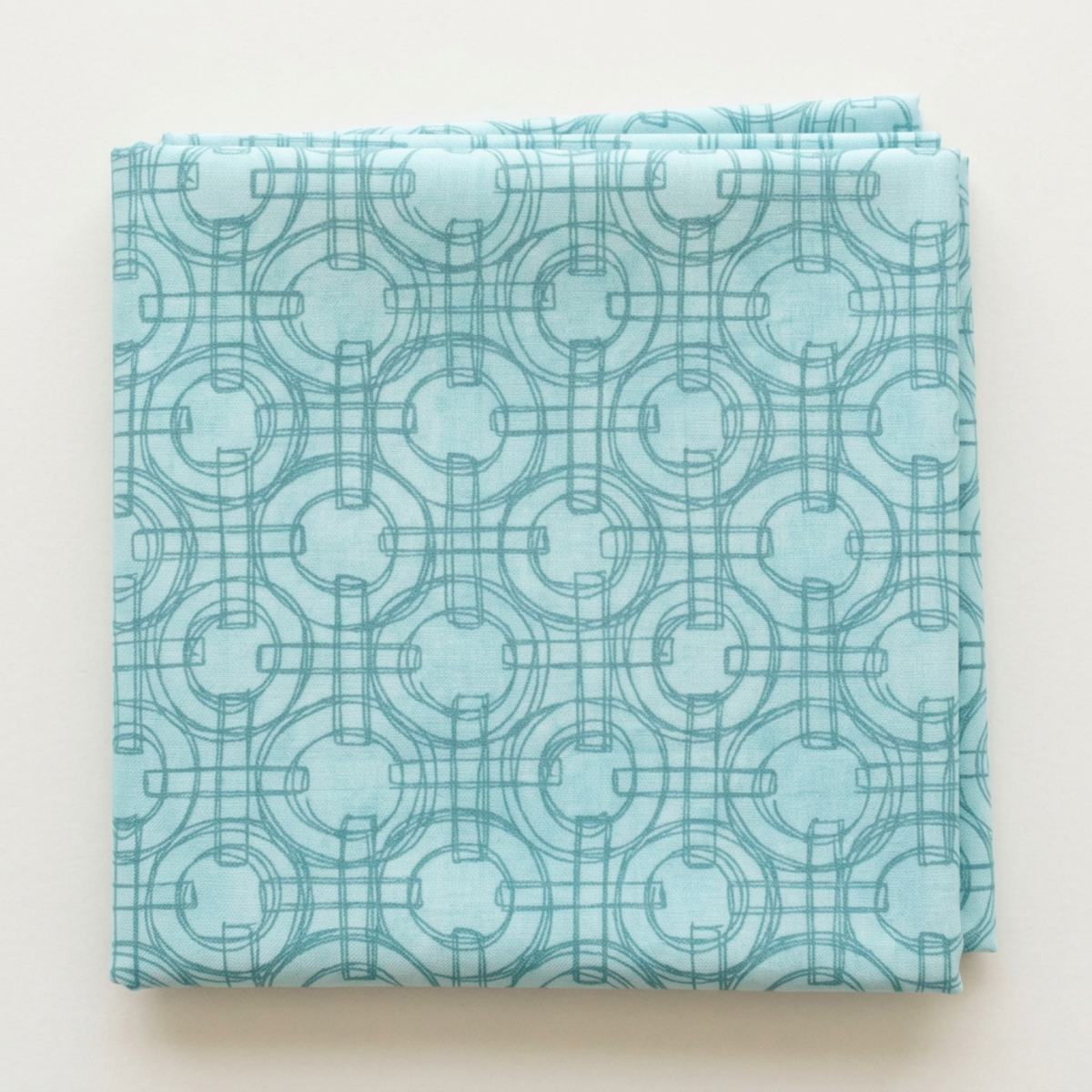 Folded Serena Shores Mist fabric by Robin Pickens for Moda Fabrics on a light surface, showing muted teal linked rings and intersecting lines on pale aqua cotton.