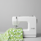 White quilting cotton with watercolor tropical banana leaves and green fern sprigs draped over a modern white sewing machine — Safari Fun Jungle Leaves White by Terry Runyan for Benartex
