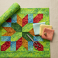 Nature-inspired jewel-tone quilt project flat lay featuring Expressions Batiks Hand-Dyes Shamrock fabric by Riley Blake Designs as the focal fabric – bright electric lime-green hand-dyed batik with chartreuse yellow and teal-blue mottled overlays in pieced blocks with coordinating forest green, tomato red, curry yellow, and periwinkle blue tonal batiks on a warm linen surface