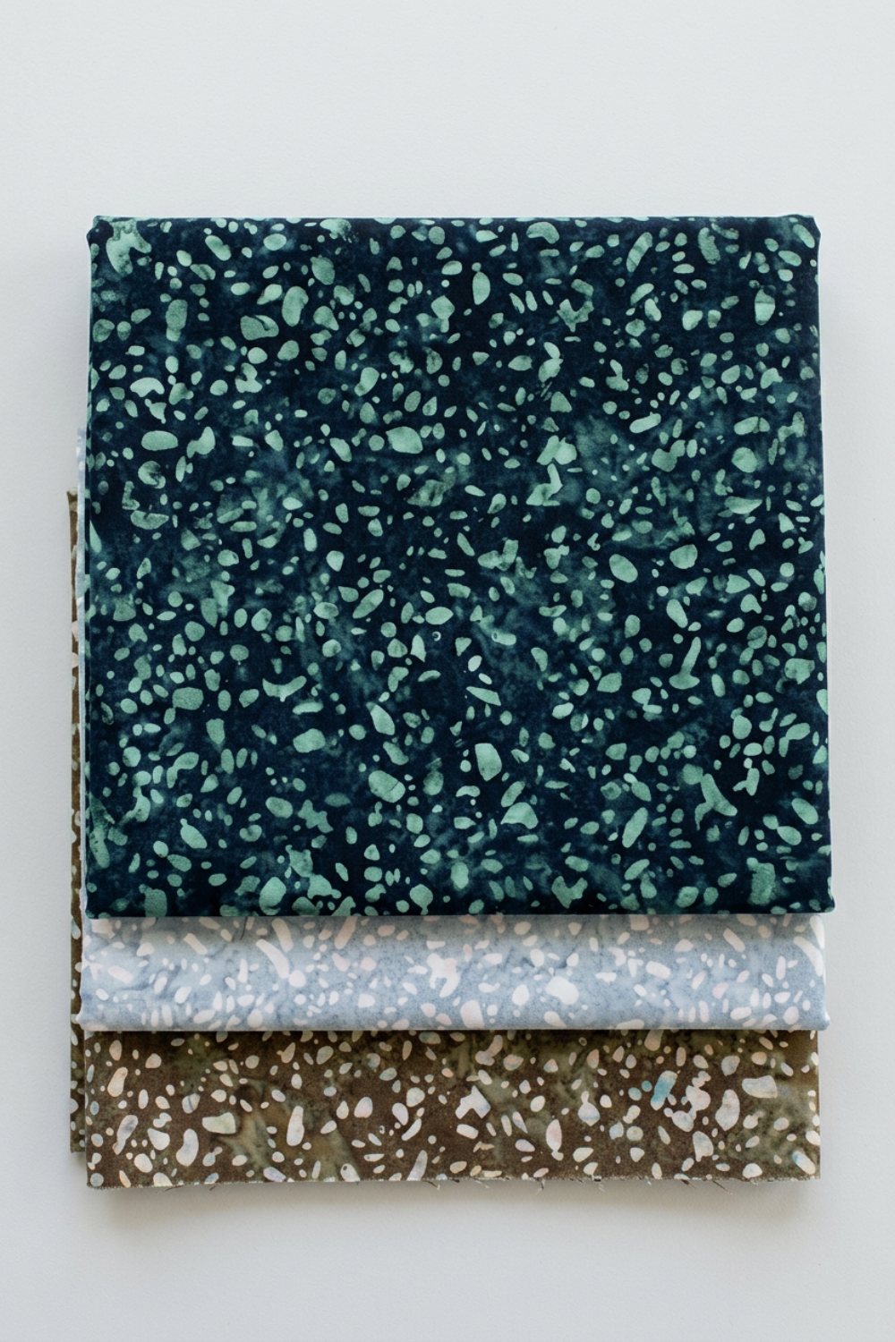 Terrazzo Smoky Teal batik fabric by Riley Blake Designs stacked above coordinating pale blue-gray and khaki speckled fabrics on a light gray background.