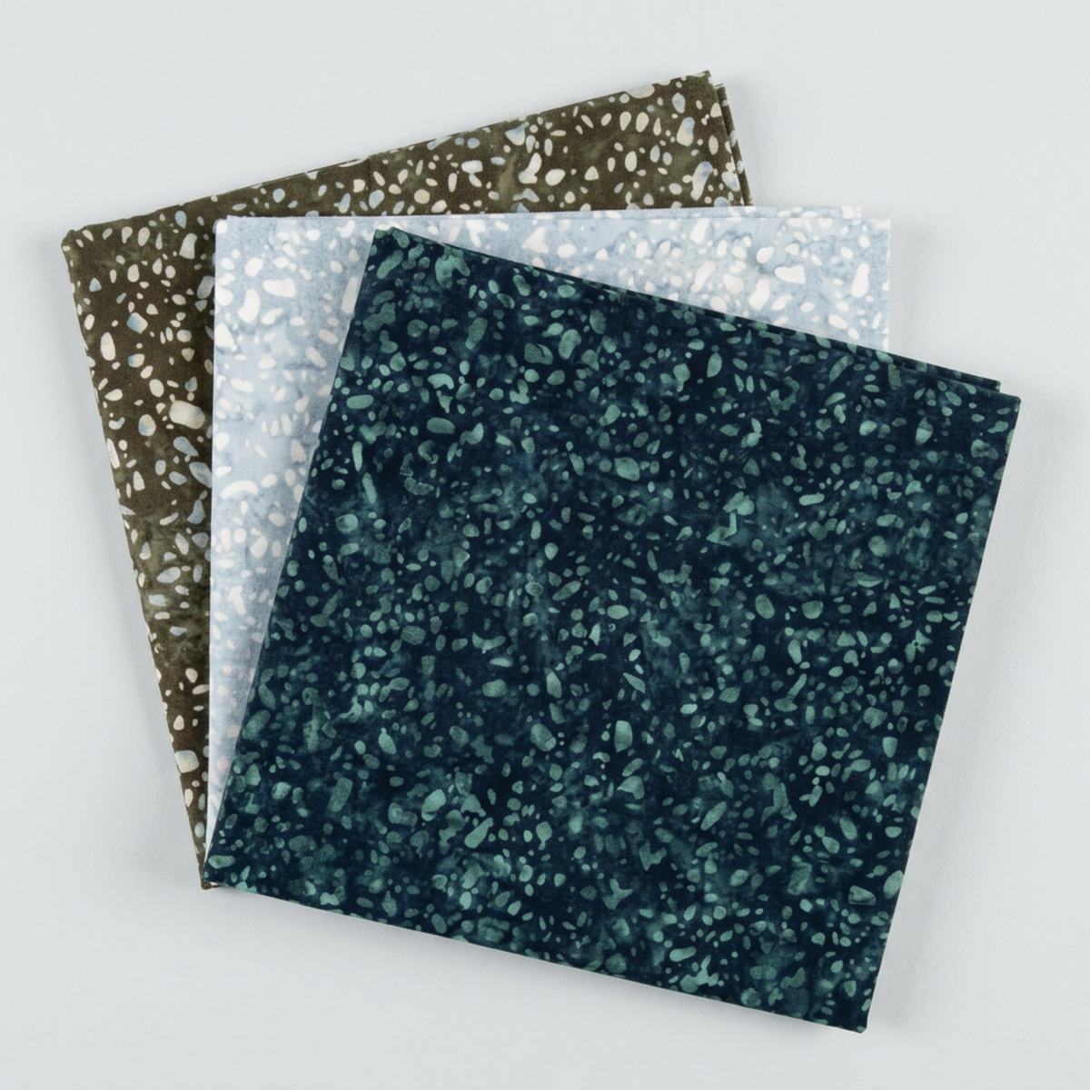 Terrazzo Smoky Teal batik fabric by Riley Blake Designs fanned in front of coordinating pale blue-gray and khaki speckled fabrics on a light gray background.