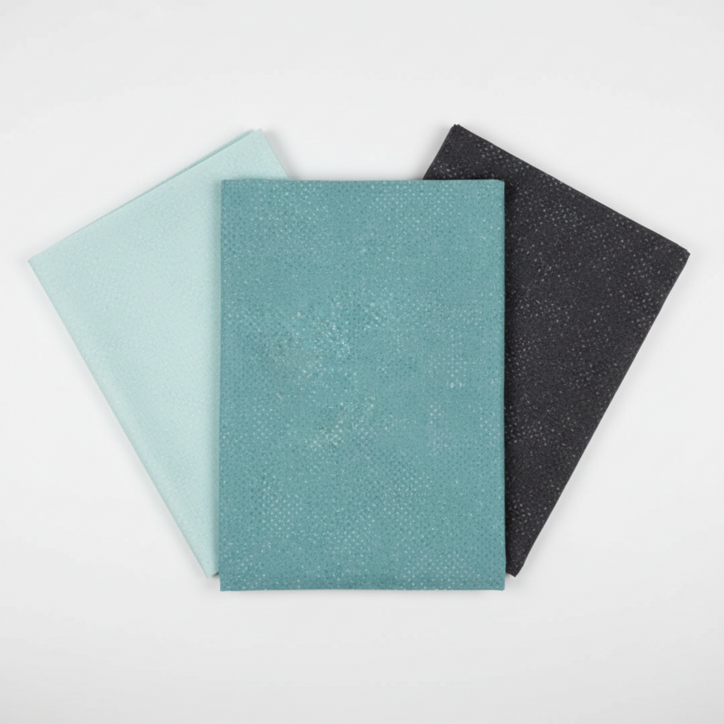 Three coordinating tonal speckled near-solid blender fabrics in a fan arrangement featuring Spotted Dusty Teal by Zen Chic for Moda Fabrics in medium teal-blue, with Spotted Mist in pale aqua-blue and Spotted Dark Slate in deep charcoal – 100% cotton
