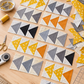 Flying geese blocks in progress for the Spotted Geese quilt, featuring Filigree Rice Saffron fabric by Zen Chic for Moda Fabrics with gray and cream coordinating fabrics, scraps, and sewing tools on a wooden table. Quilts shown for inspiration feature the Spotted Geese Quilt Pattern by Zen Chic — all available at IraRobi Quilts.