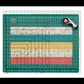Creative Grids Stripology XL Ruler laid flat on a green self-healing cutting mat over alternating coral, teal, yellow, and cream quilting fabric strips visible through the clear acrylic, with a rotary cutter at the top right corner