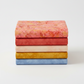 Expressions Batiks Hand-Dyes Sunset fabric by Riley Blake Designs on top of a coordinating stack of five tonal batik fabrics – soft coral-pink on top with tomato red, soft peach-cream, curry yellow, and periwinkle blue tonal batiks below, 100% cotton