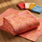 Expressions Batiks Hand-Dyes Sunset fabric by Riley Blake Designs draped on a warm wood surface with coordinating tomato red, soft peach-cream, curry yellow, and periwinkle blue tonal batik fabric pieces nearby – soft coral-pink hand-dyed batik with warm golden-orange splashes, soft peach tones, and hints of deeper rose, a luminous warm focal blender perfect for warm-toned and floral quilts, 100% cotton batik