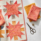 Warm-toned floral quilt project flat lay featuring Expressions Batiks Hand-Dyes Sunset fabric by Riley Blake Designs as the focal fabric – soft coral-pink hand-dyed batik with warm golden-orange splashes and peach tones in pieced blocks with coordinating tomato red, peach-cream, curry yellow, and periwinkle blue tonal batiks on a soft white linen surface