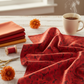 Sunshine Red Daisies 3 Yard Cut quilting cotton by Jason Yenter for In The Beginning Fabrics — loosely draped on a white wooden surface with coordinating warm red, burnt orange, and gold fabric swatches, red thread spool, gold scissors, dried orange dahlias, and a mug of tea in a cozy sewing room. Quilt blocks shown for inspiration. Coordinating fabrics and quilt patterns are available at IraRobi Quilts.