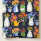 Close-up of Superbloom Flea Market Vases Indigo fabric by Robin Pickens for Moda Fabrics, showing floral pitchers, green jars, yellow watering cans, and butterflies, with a soft fold along the lower edge.