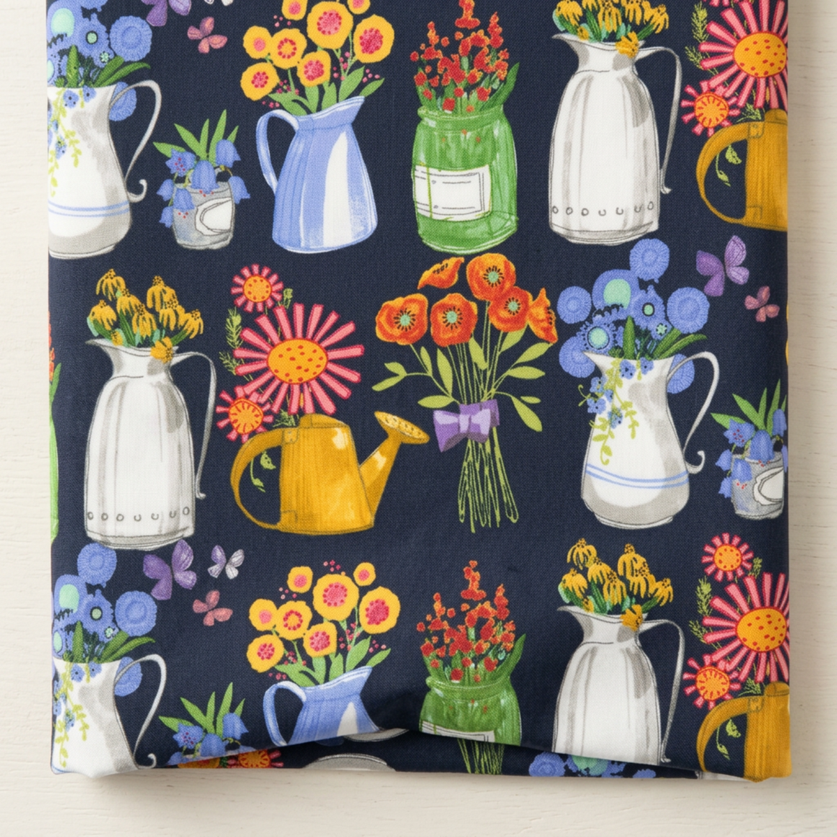 Close-up of Superbloom Flea Market Vases Indigo fabric by Robin Pickens for Moda Fabrics, showing floral pitchers, green jars, yellow watering cans, and butterflies, with a soft fold along the lower edge.