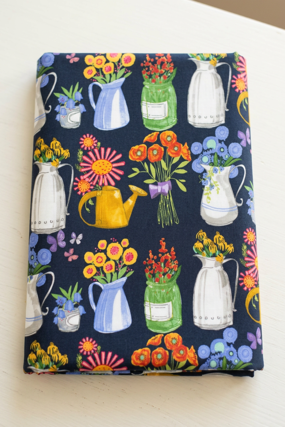 Neatly folded Superbloom Flea Market Vases Indigo fabric by Robin Pickens for Moda Fabrics on a light tabletop, showing colorful flower-filled containers and butterflies on dark indigo cotton.