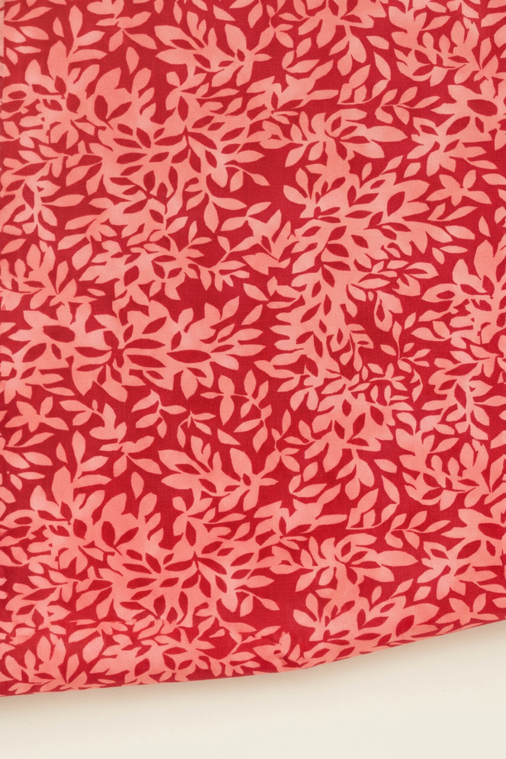 Close-up of Superbloom Leafy Geranium fabric by Robin Pickens for Moda Fabrics, showing dense coral-pink leafy branches on rich red cotton with a gently curved lower edge against a light surface.