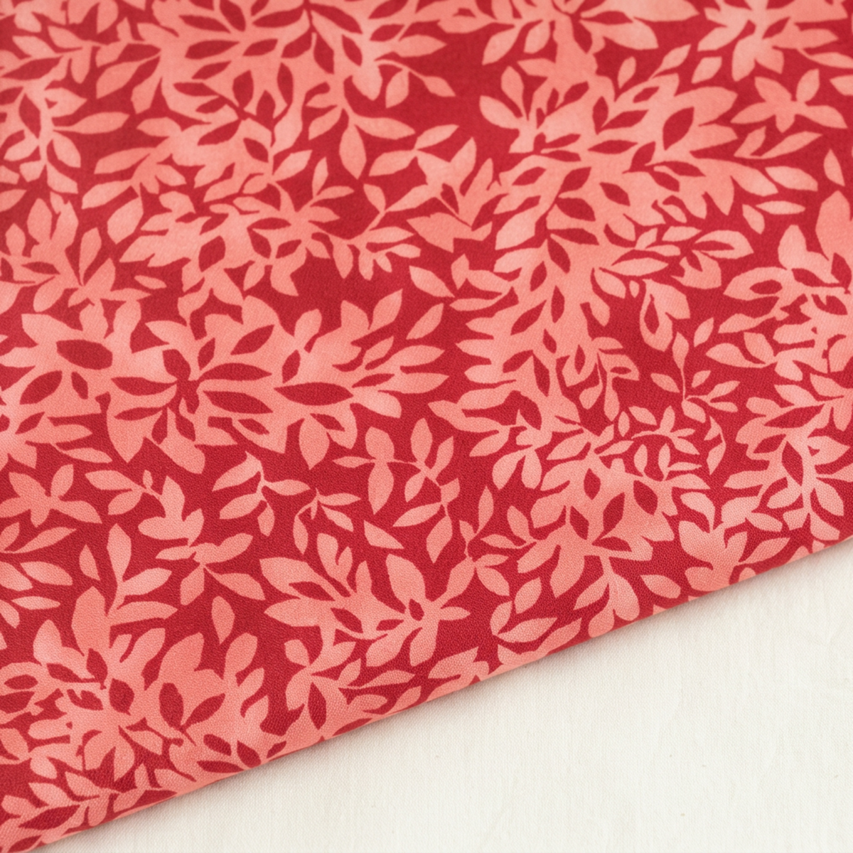 Close-up of Superbloom Leafy Geranium fabric by Robin Pickens for Moda Fabrics, showing coral-pink leaves on rich red cotton with a diagonal fabric edge against a light surface.