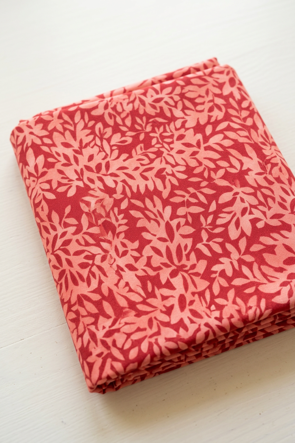 Superbloom Leafy Geranium fabric by Robin Pickens for Moda Fabrics, neatly folded at an angle on a light tabletop, showing coral-pink leaves on geranium-red cotton.