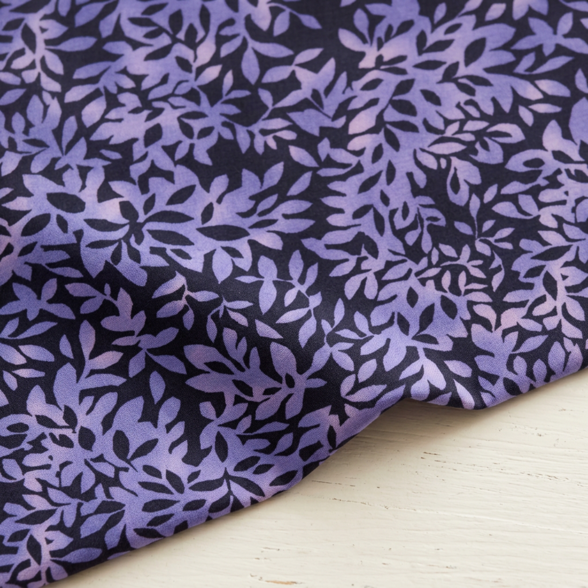 Close-up of Superbloom Leafy Indigo fabric by Robin Pickens for Moda Fabrics, softly draped on a light tabletop with lavender-purple leaves on a dark indigo ground.