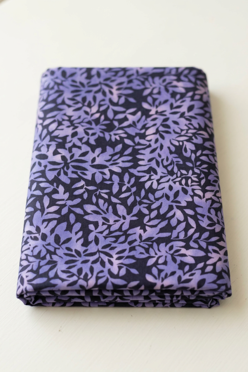 Neatly folded Superbloom Leafy Indigo fabric by Robin Pickens for Moda Fabrics on a light tabletop, showing lavender-purple leaves on dark indigo cotton.