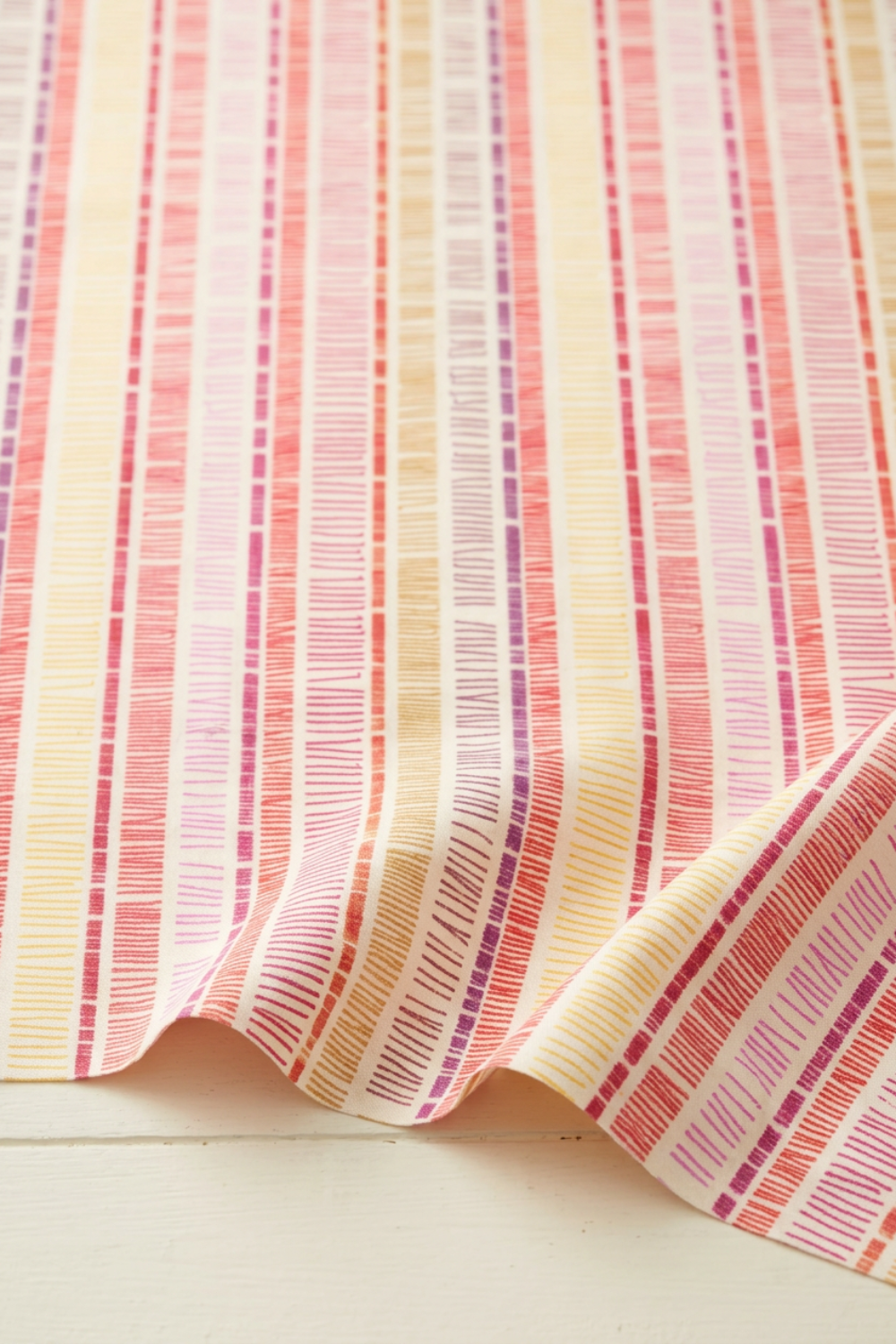 Superbloom Scribble Stripe Cream fabric by Robin Pickens for Moda Fabrics, softly draped in folds along the lower edge, showing coral-red, pink, plum, and golden-yellow stripes on cream cotton.