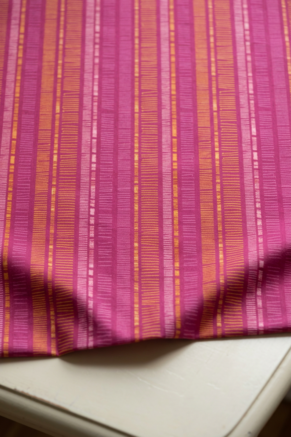 Superbloom Scribble Stripe Orchid fabric by Robin Pickens for Moda Fabrics, softly draped along a tabletop edge, showing golden-orange and pink scribble stripes on orchid-magenta cotton.