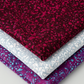 Terrazzo Mahogany batik fabric by Riley Blake Designs stacked above coordinating pale blue-gray and aqua-speckled purple fabrics on a light background.