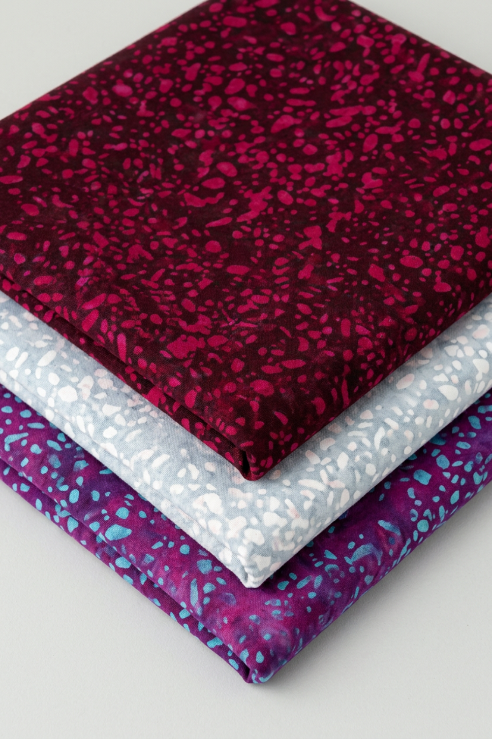 Terrazzo Mahogany batik fabric by Riley Blake Designs stacked above coordinating pale blue-gray and aqua-speckled purple fabrics on a light background.