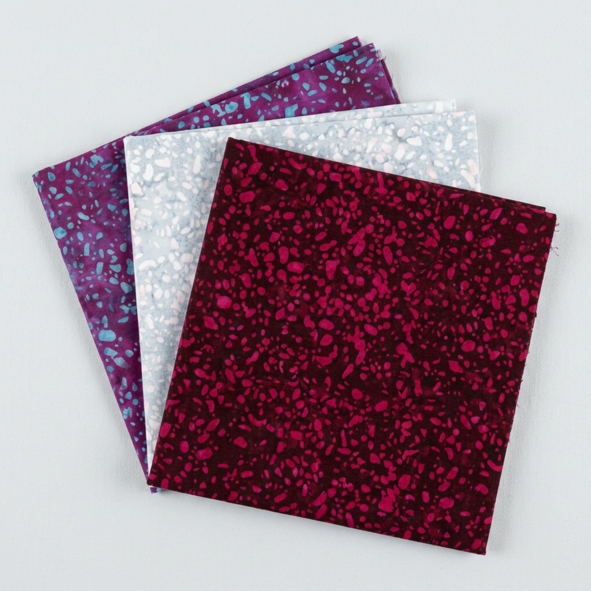 Terrazzo Mahogany batik fabric by Riley Blake Designs fanned in front of coordinating pale blue-gray and aqua-speckled purple fabrics on a light gray background.