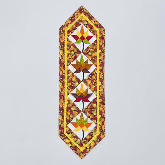 Vermont Foliage handmade quilted table runner with four autumn-colored maple leaf blocks on white diamonds, curved black stems, a yellow inner border and pointed ends on light gray.