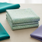 Vivid Mums Spice Teal fabric by Jason Yenter for In The Beginning Fabrics folded in a stack on a light linen surface with coordinating teal, emerald, and periwinkle fabrics – dense allover chrysanthemum burst blender with tiny pointed petals in teal, emerald, and sage on light cream and silver-grey, perfect for teal and jewel-toned quilt projects, 100% cotton, 3 yard cut