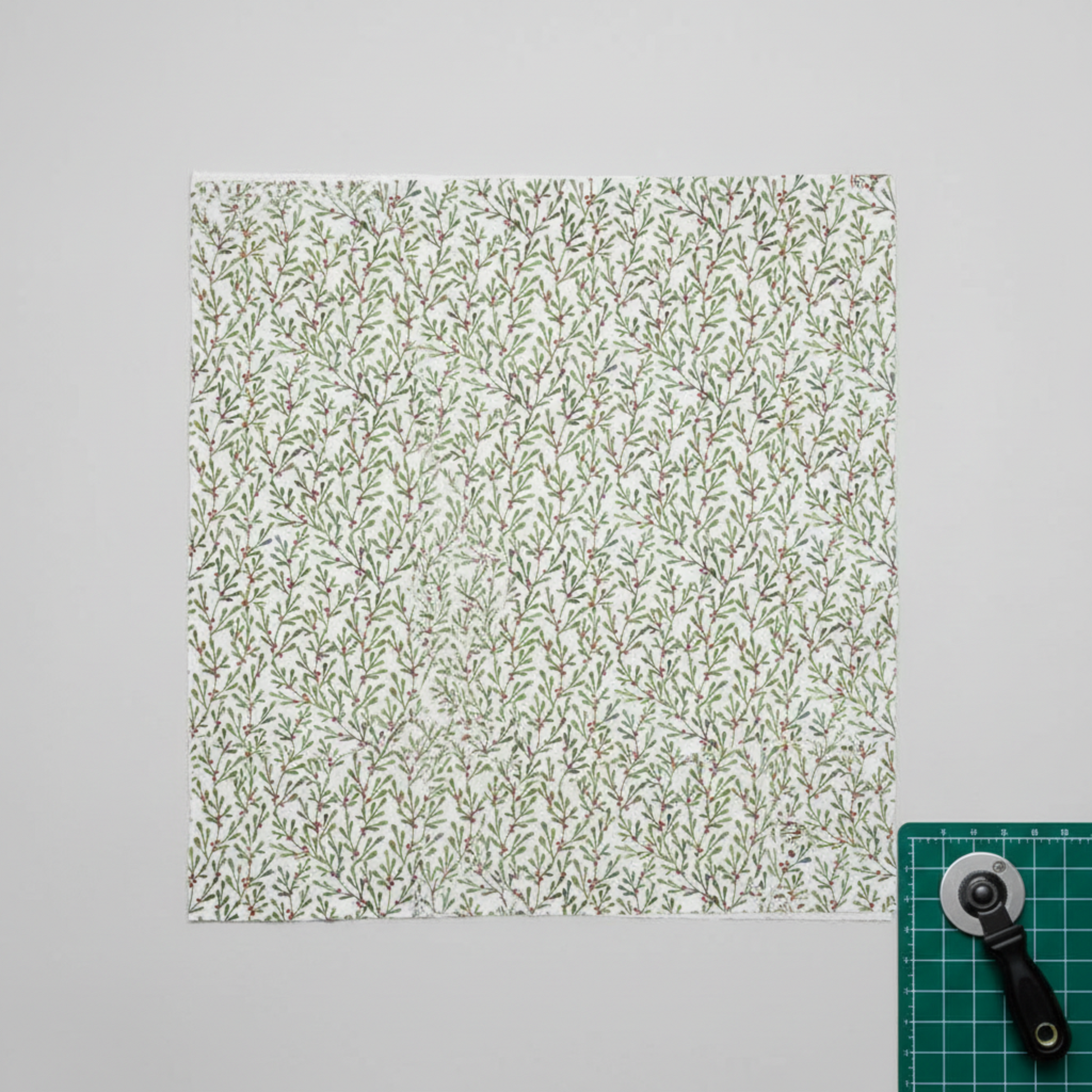 Winter Solstice II White Branches quilting cotton by Jason Yenter for In The Beginning Fabrics — fabric laid flat showing allover watercolor mistletoe sprigs in soft sage and forest green with tiny red berries on crisp white, shown in a clean modern quilting workspace with cutting mat and rotary cutter, 100% cotton, 43–44" wide. Quilt blocks shown for inspiration. Coordinating fabrics and quilt patterns are available at IraRobi Quilts.