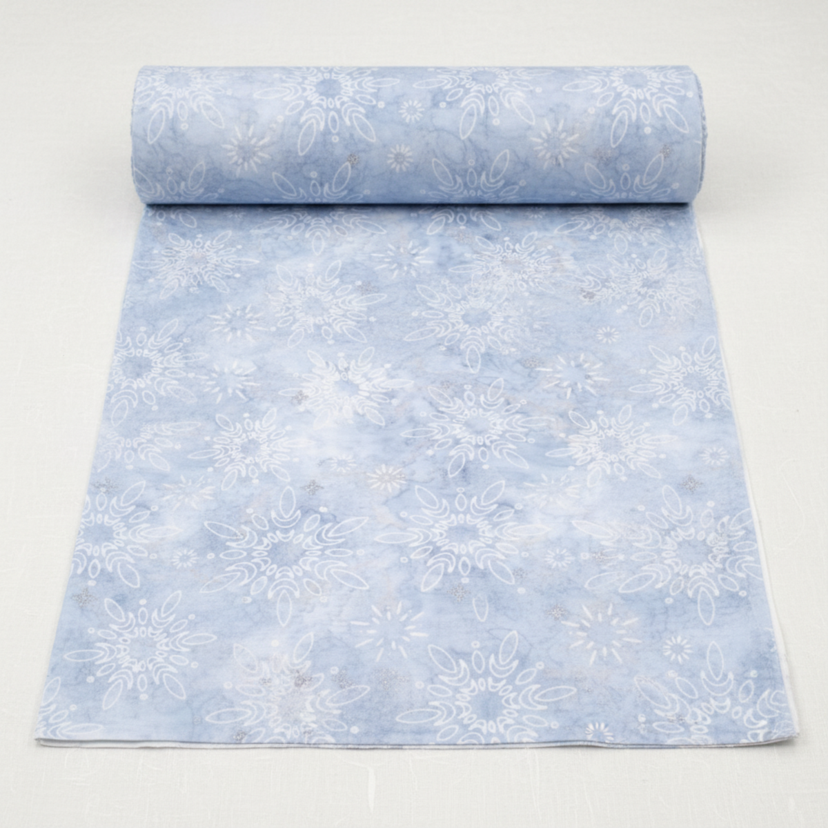 Winter Sparkle Glacier Artisan Batiks quilting cotton by Robert Kaufman Fabrics shown as a partially unrolled bolt with a cut yard of fabric displaying the full print repeat — soft powder blue tone-on-tone batik with white snowflake and daisy medallion motifs and silver metallic glitter accents.