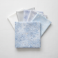 Winter Sparkle Glacier Artisan Batiks by Robert Kaufman Fabrics shown in a coordinating fabric stack with crisp white, soft silver-gray, pale periwinkle batik, and white-on-white snowflake print companions. Quilt blocks shown for inspiration. Coordinating fabrics and quilt patterns are available at IraRobi Quilts.