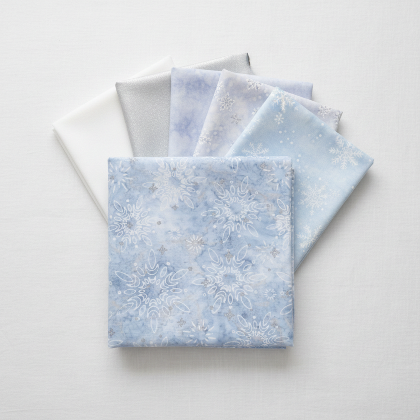 Winter Sparkle Glacier Artisan Batiks by Robert Kaufman Fabrics shown in a coordinating fabric stack with crisp white, soft silver-gray, pale periwinkle batik, and white-on-white snowflake print companions. Quilt blocks shown for inspiration. Coordinating fabrics and quilt patterns are available at IraRobi Quilts.