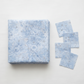 Winter Sparkle Glacier Artisan Batiks quilting cotton by Robert Kaufman Fabrics – folded fabric stack showing soft powder blue tone-on-tone batik with white snowflake and daisy medallion motifs and silver metallic glitter accents. Quilt blocks shown for inspiration. Coordinating fabrics and quilt patterns are available at IraRobi Quilts.