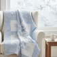 Cozy winter quilt draped over a white armchair featuring Winter Sparkle Glacier Artisan Batiks by Robert Kaufman Fabrics in soft powder blue with white snowflake motifs and silver metallic accents. Quilt blocks shown for inspiration. Coordinating fabrics and quilt patterns are available at IraRobi Quilts.