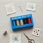 WonderFil Inspira Specialty Bobbin Thread blue box with 5 metallic spools on a linen surface, surrounded by fabric swatches showing gold, turquoise, and red metallic embroidery samples, a hand-sewing needle, scissors, and a silver thimble
