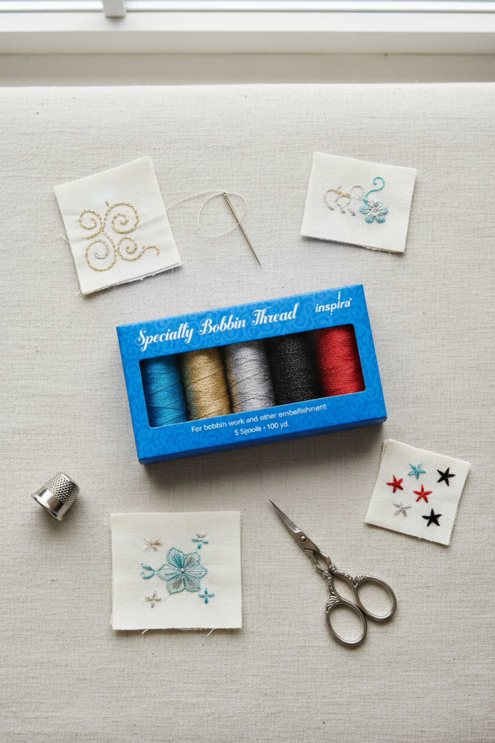 WonderFil Inspira Specialty Bobbin Thread blue box with 5 metallic spools on a linen surface, surrounded by fabric swatches showing gold, turquoise, and red metallic embroidery samples, a hand-sewing needle, scissors, and a silver thimble