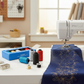 WonderFil Inspira Specialty Bobbin Thread blue box and metallic spools in turquoise, silver, black, and red on a white sewing table beside a sewing machine stitching a gold and silver metallic floral design onto navy blue fabric, with bobbins and a needle case nearby in a bright studio