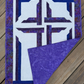 The Wondrous Cross wall quilt on wooden decking, with the lower-right corner folded back to show purple patterned backing, binding, and quilting stitches.