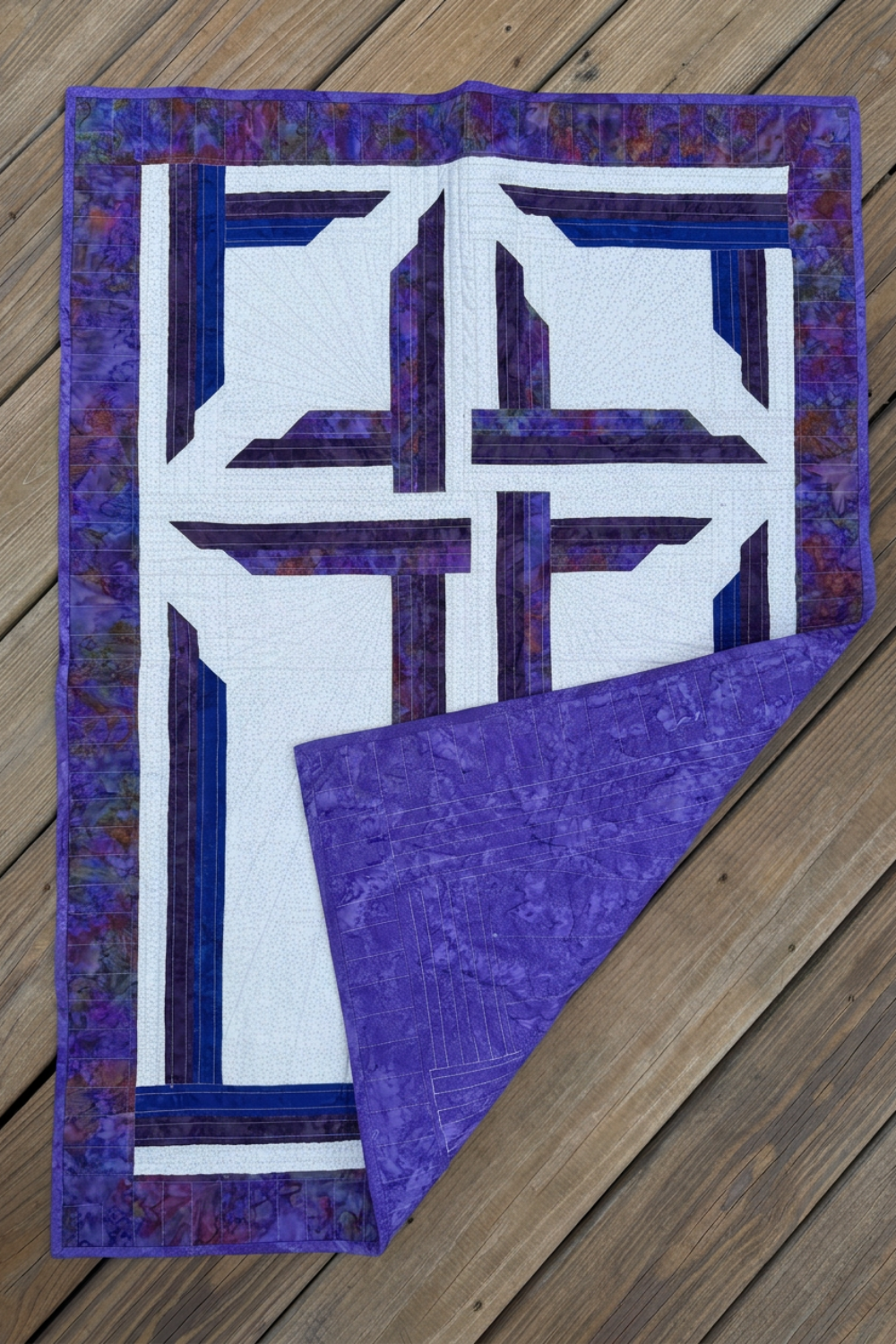 The Wondrous Cross wall quilt on wooden decking, with the lower-right corner folded back to show purple patterned backing, binding, and quilting stitches.