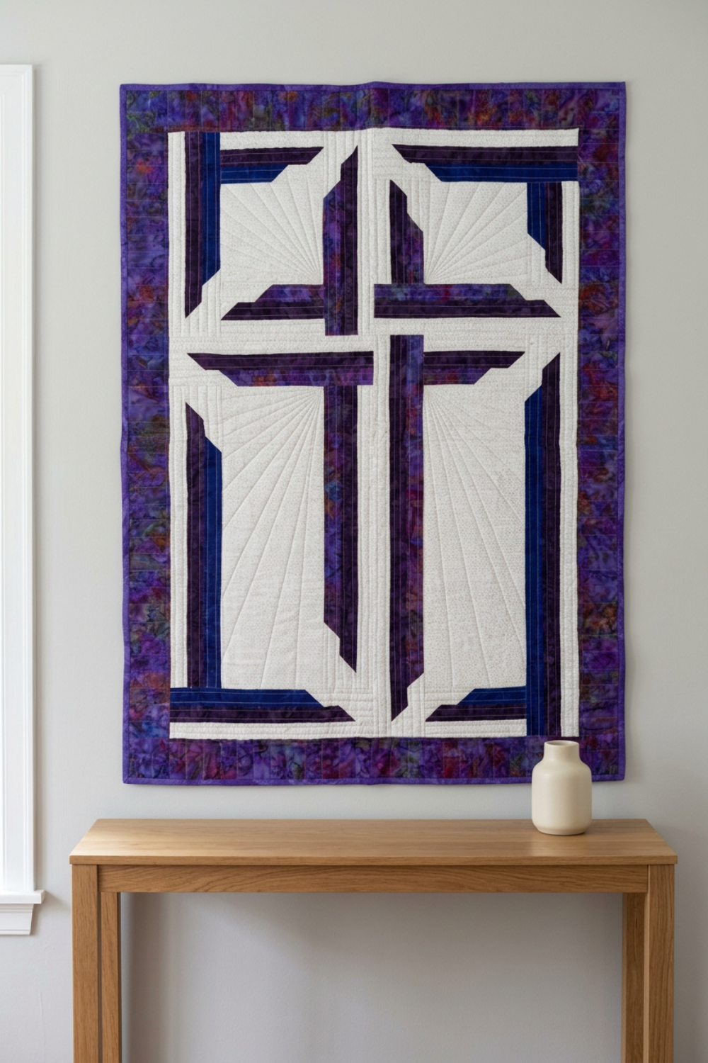 The Wondrous Cross purple, blue, and white wall quilt hanging above an oak console with a cream vase beside a window.