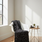 Writing on the Wall Script Black quilting cotton by FreeSpirit Fabrics -- bold black script fabric draped over a white modern chair in a bright loft sewing studio near a large window, with thread spools, scissors, and ceramic mug on a side table
