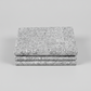 Writing on the Wall Well Worded White quilting cotton by FreeSpirit Fabrics -- three folded yards stacked on a grey surface, grey and white ground with white outlined words in varying sizes and directions including special, beauty, serendipity, magic, imagination, harmony, wow, and tomorrow